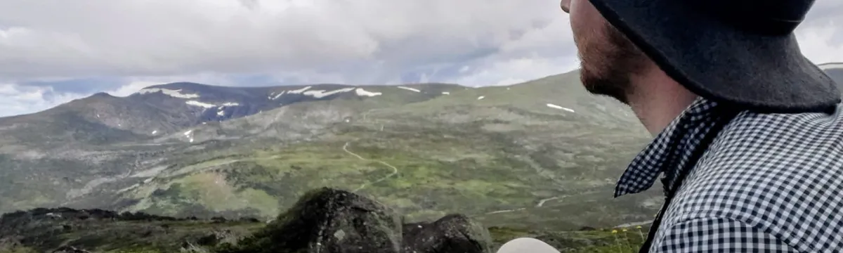 Josh sits on a grassy alpine slope, facing away from the camera and looking out over a mountainous landscape with patches of snow and cloud-covered skies. They wear a wide-brimmed dark hat, a checkered long-sleeve shirt, and light-coloured pants. In the foreground, native alpine plants with tall white flower stalks are visible, adding texture to the natural scene