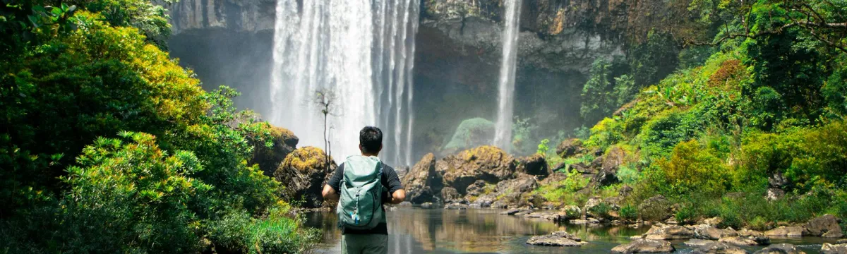 A person wearing a green backpack stands on a rock in the middle of a shallow river, facing a large waterfall that cascades from a tall cliff surrounded by lush, tropical forest. The individual appears to be hiking or exploring, dressed in outdoor gear including a short-sleeved shirt, shorts, and socks with sneakers. Bright sunlight filters through the trees, highlighting the greenery and the mist from the waterfall.
