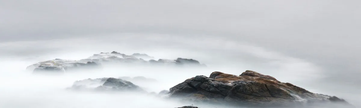Coastal rocks appearing out of mist on the ocean. The sky is grey.