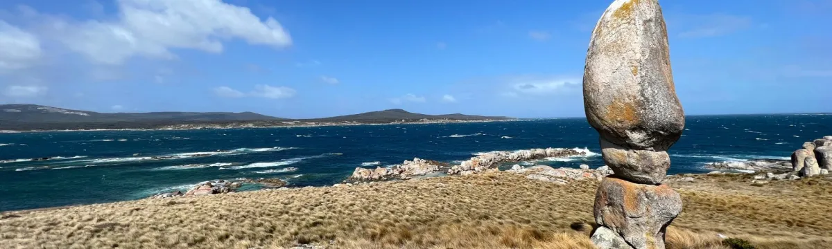 A coastal landscape featuring a strikingly tall and narrow balanced granite boulder standing upright on grassy terrain in the foreground. The scene overlooks a windswept ocean with white-capped waves and rocky outcrops. In the distance, rolling hills stretch across the horizon beneath a blue sky dotted with clouds. The location has a wild, remote feel.