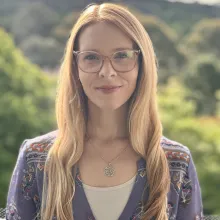 A picture of Dr Elle Bowd. She wears framed glasses, and has long gold-blonde hair. Behind her head and shoulders is a blurred bushland landscape.