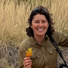 Researcher, Ellen Ryan-Colton, kneels in a patch of dry grass holding a grevillea flower.