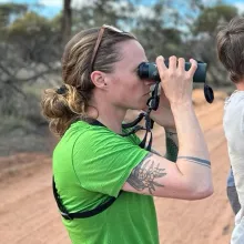 Laura Bussolini holds up binoculars to her eyes. She is wearing a green shirt and standing on a dusty road.