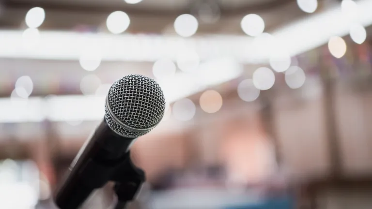 A silver microphone faces the viewer while the background is blurred, bright, with lots of lights.