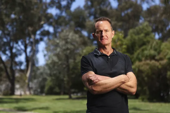 Andrew Macintosh wears a black polo shirt and stands with his arms crossed facing the camera in front of a trees and grass