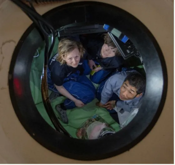Three people wearing work clothing sit closely together in a small, enclosed space viewed from above through a circular hatch or window. They are looking upward toward the camera. The setting appears industrial or scientific, possibly a confined research or training module. The floor is padded with green material, and wires and equipment are visible around the edge of the opening