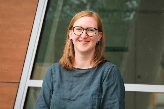 Bec Colvin looks at the camera, smiling. She is wearing black glasses and a dark blue/grey shirt.