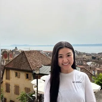 A portrait photo of Shengnan Wang who smiles at the camera in the foreground. In the background is a port city.