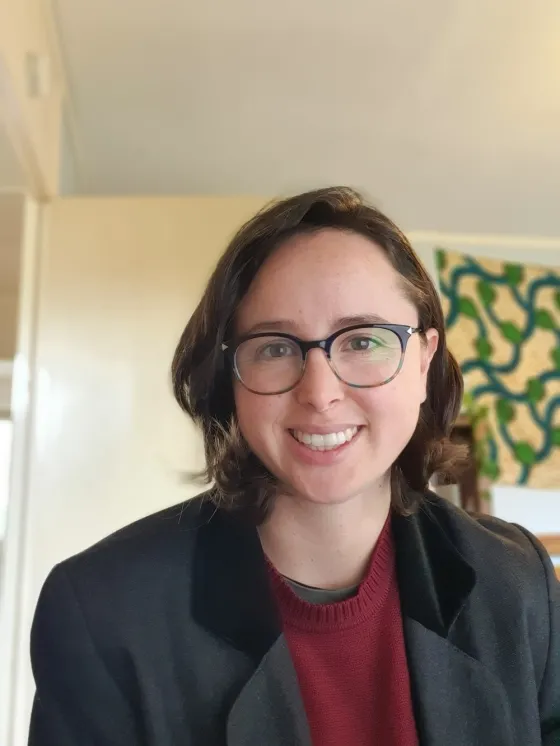 Carla Alexandra smiles at the camera, she has short brown hair and glasses. The photo is taken indoors, and Carla is facing the camera. They are wearing a dark blazer over a maroon sweater. The background shows a softly lit room with cream-colored walls and a piece of art or fabric with green and blue patterns hanging on the wall. Natural light brightens the space, giving the image a warm and welcoming feel.