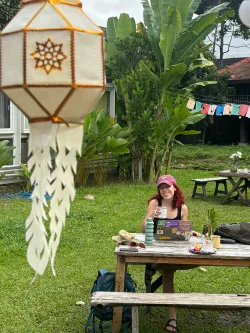 Isabelle sits at a timber table outside with a cup of tea in her hand. There are banana trees in the background, colourful bunting and a hut-like building. There is a lantern in the foreground.