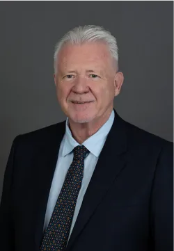 David Brand looks and smiles at the camera. He wears a pale blue shirt, black suit jacket and black spotted tie.