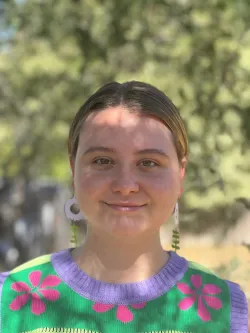 Tegan Clark smiles at the camera. She wears a green vest with pink flowers and purple neck.