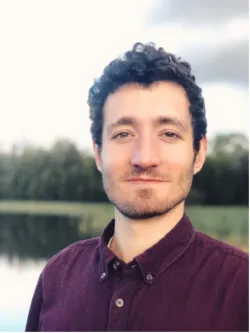 A head-and-shoulders portrait of Martino Malerba standing outdoors near a body of water bordered by trees. He has short, dark curly hair and a closely trimmed beard. He’s wearing a button-up shirt in a deep burgundy colour. The background is softly blurred, with greenery and sky visible behind him.