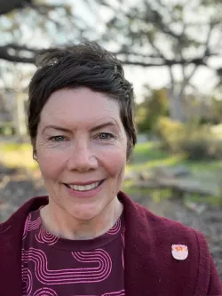 A profile photo of Rosie Cooney, who is smiling at the camera in an outdoor setting with dappled sunlight and trees in the background. She is wearing a patterned burgundy top and a maroon jacket with a small enamel badge pinned to the lapel. The background is softly blurred, drawing attention to her face, and the overall mood is warm and natural.