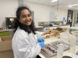 Sharmin is working in a laboratory, she wears a white lab coat and blue gloves.