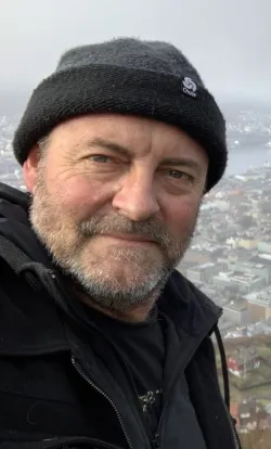 A profile photo of Simon Haberle, wearing a black knit beanie and a zip-up black jacket smiles slightly at the camera in a close-up selfie. In the background, a misty cityscape with clustered buildings and a harbor is visible below