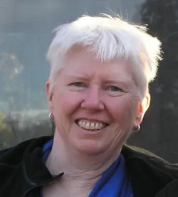 Sue McIntyre smiles at the camera. She is wearing a blue top and black jacket. They are looking directly at the camera in a warmly lit outdoor setting, with a blurred background of trees and sky behind them.