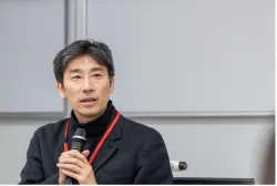 Taro holds a microphone in his hands. He has black hair and is wearing a black jacket.