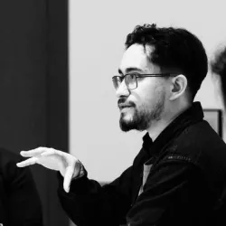A black and white portrait of Adrian. He has short dark hair and a neatly trimmed beard, wearing glasses and a dark jacket. He is indoors and captured mid-gesture, as if speaking or explaining something.