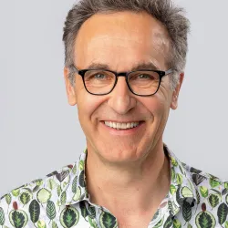 A photo of a man looking at the camera with black glasses and a green shirt with trees on it.