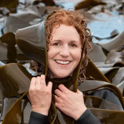 Researcher Crid poses for a photo while wrapped up in giant kelp. She is smiling and facing the camera.