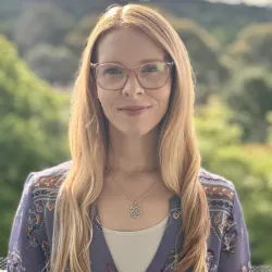 A picture of Dr Elle Bowd. She wears framed glasses, and has long gold-blonde hair. Behind her head and shoulders is a blurred bushland landscape.