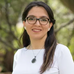 Fateme stands in a white shirt with black glasses on, outside near a tree.