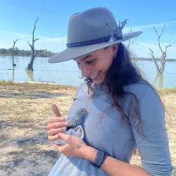 Grace Hodder holds a baby parrot in her hands. There is a small lake behind her and she wears an Akubra hat.