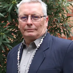 Associate Professor Ian Fry stands in front of a native shrub at the Fenner School and looks at the camera. He is wearing a suit and a string of shells around his neck.