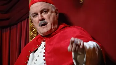 A close-up of actor John Cleese dressed in Roman papal attire, and pointing at the camera while mid-speech.