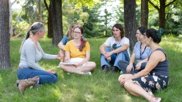Students sit in a half circle on a grassy knoll while listening to a lecturer speak.