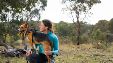 A Fenner researcher, Shoshana Rapley, kneels with her dog on her leg and looks at her, with woodland in the background.