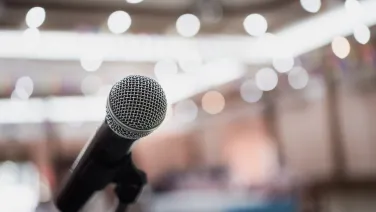 A silver microphone faces the viewer while the background is blurred, bright, with lots of lights.