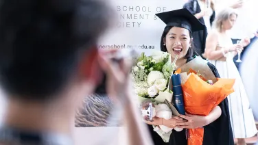 A Fenner graduate stands in front of a banner and smiles while people in the foreground take photos.