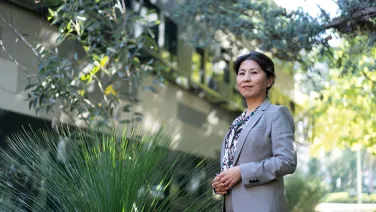 Researcher, Xuemei Bai, stands in front of the Fenner building with a grass tree beside her.