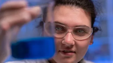 A student holds a beaker of blue liquid in front of her face. She is wearing clear goggles.