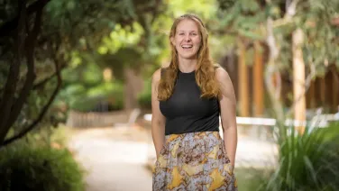 Student, Isobel, stands in a leafy part of the campus smiling and facing the camera.