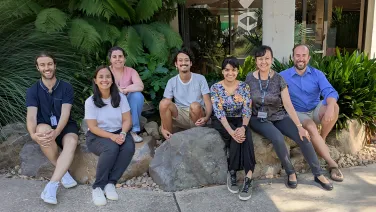 A group of researchers sit among large rocks and ferns outside a building.