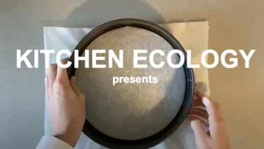 On a direct angle, the circular shape of a cake tin sits against a light grey kitchen counter. There is baking paper in the middle of the circle. Over the top of this symmetrical image are the words 'Kitchen Ecology'