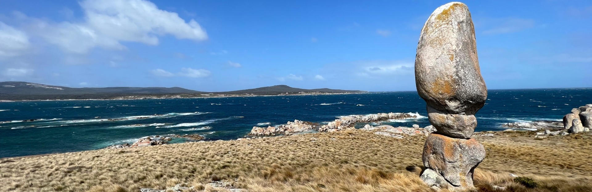 A coastal landscape featuring a strikingly tall and narrow balanced granite boulder standing upright on grassy terrain in the foreground. The scene overlooks a windswept ocean with white-capped waves and rocky outcrops. In the distance, rolling hills stretch across the horizon beneath a blue sky dotted with clouds. The location has a wild, remote feel.